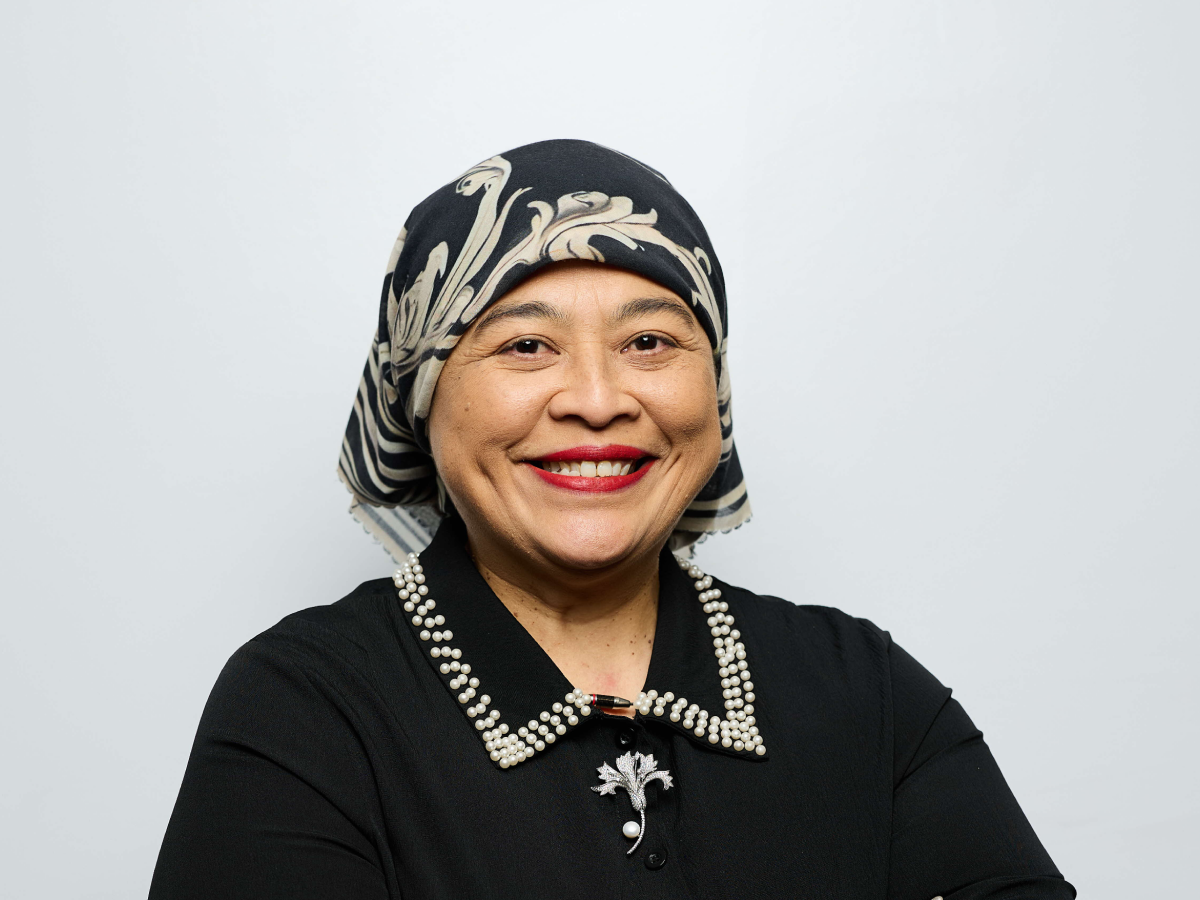 Mdm A J Suhani in a black outfit and patterned headscarf smiling with arms crossed against a light grey background